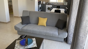 Un salon avec un canapé gris, une table basse sur laquelle se trouve un objet bleu, et une cuisine en arrière-plan.