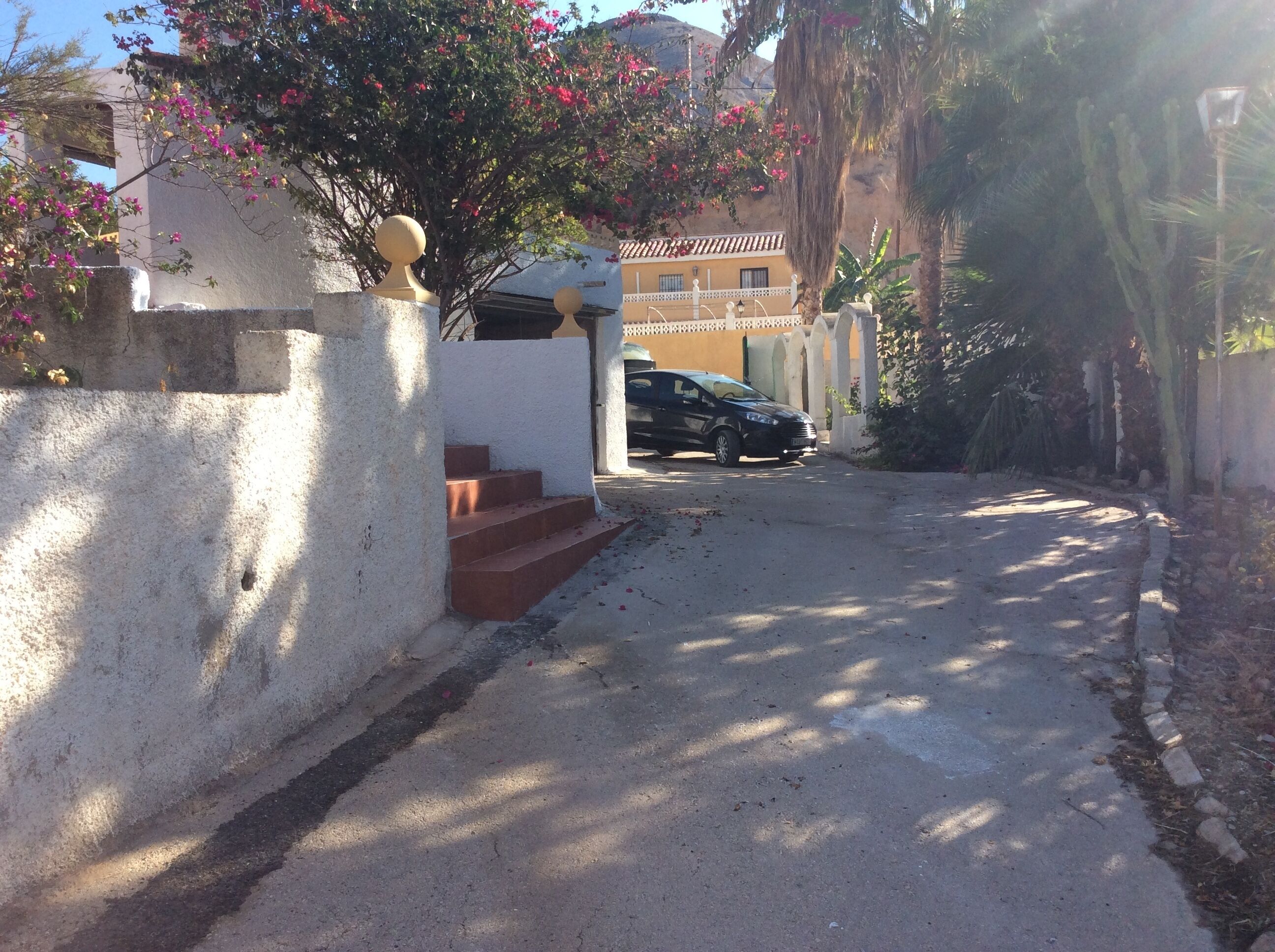 A narrow street with a parked car, a white building with a balcony, and a tree with pink flowers.