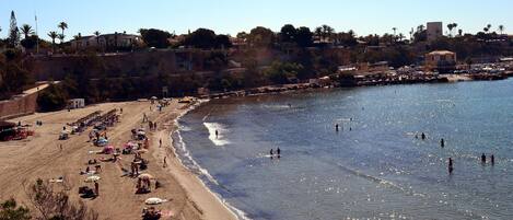 Una spiaggia con persone che prendono il sole e nuotano, una riva sabbiosa e un promontorio roccioso con edifici.
