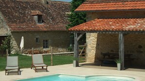 Une piscine avec des chaises longues, un bâtiment en pierre doté d’un toit en tuiles et un espace salon abrité.