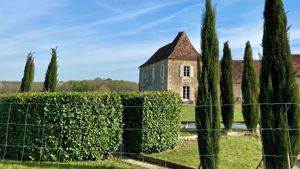 Un bâtiment en pierre surmonté d’un toit triangulaire, entouré de grands arbres élancés et d’un arbuste taillé avec soin.
