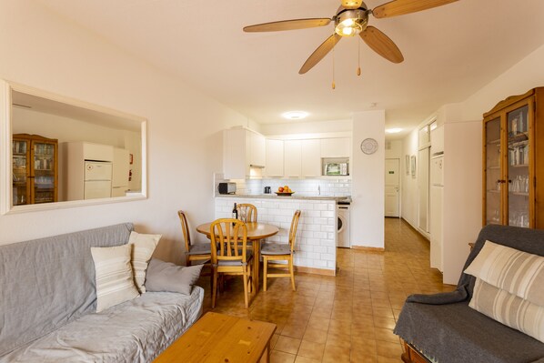A living room with a sofa, a coffee table, and a dining table with chairs. There is a kitchen area with a refrigerator, a microwave, and a sink. A ceiling fan is visible above the dining area. - Costa Adeje ground floor 1 bedroom sea & mountain view apartment (Costa Adeje)