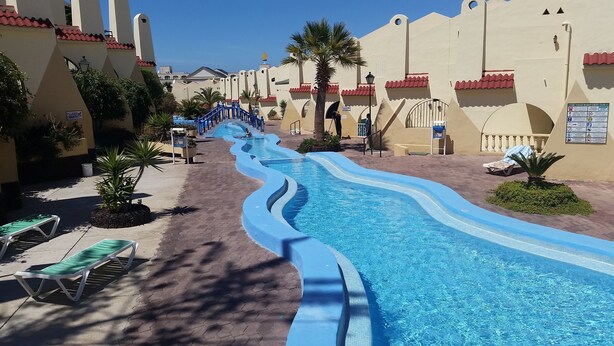 Una piscina con un diseño serpenteante, rodeada de palmeras y tumbonas, con edificios de tejas rojas al fondo.