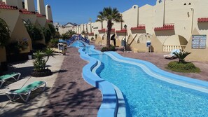 Una piscina con un diseño serpenteante, rodeada de palmeras y tumbonas, con edificios de tejas rojas al fondo.