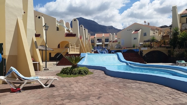 A resort pool area with a water slide, lounge chairs, and a mountain backdrop.