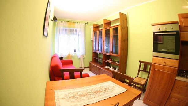 A living room with a wooden dining table, red sofa, and a kitchen area with wooden cabinets and an oven. - MAISON CASSIUS LO VERT (ISSOGNE  ITALIA)