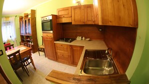 A compact kitchen with wooden cabinets, a sink, a stove, and a dining area with chairs and a table. - MAISON CASSIUS LO VERT (ISSOGNE  ITALIA)