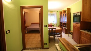 A compact kitchen with wooden cabinets, a dining area with a table and chairs, and a small bedroom with a bed and a nightstand. - MAISON CASSIUS LO VERT (ISSOGNE  ITALIA)