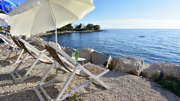 Un entorno frente a la playa con sillones de madera y una sombrilla blanca, con vistas a un mar tranquilo y a una costa rocosa.