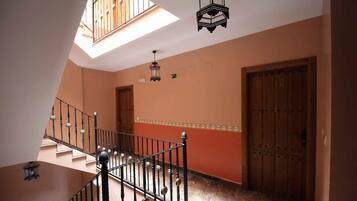 Un escalier avec une rampe métallique mène à un palier. Il y a deux portes sur le palier, dont une porte au-dessus de laquelle est inscrit le chiffre « 2 ». Les murs sont peints dans une couleur chaude, et une lucarne se trouve au-dessus de l’escalier.