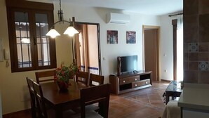 Uma área de refeições com mesa e cadeiras de madeira, uma televisão em um suporte e um vaso com planta.