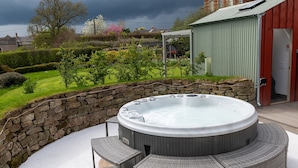 A hot tub with a stone wall and wicker furniture, surrounded by a garden and a red building in the background. - A spacious and comfortable cottage (Greenlaw)