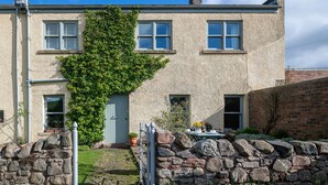 A stone house with a green ivy-covered wall, a stone wall in front, and a small garden area with a table and chairs. - A spacious and comfortable cottage (Greenlaw)