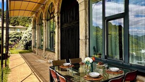 An outdoor dining area with a glass table, red chairs, and a covered patio with stone arches and large windows. - L'Orangerie • Architect house -Private pool-Nature-Village view-Calm-Rivers.  (Joannas)