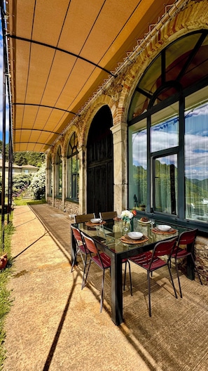 An outdoor dining area with a glass table, red chairs, and a covered patio with stone arches and large windows.