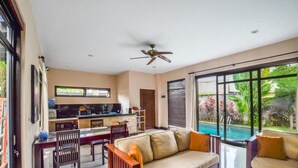 Amplia sala de estar con sofá, mesa de comedor y sillas, zona de cocina y un ventanal con vistas a una piscina.