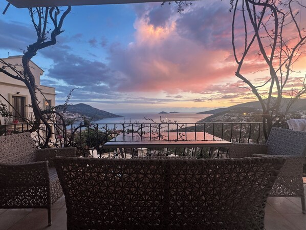 A terrace with a view of the sea and mountains, a table, and chairs.