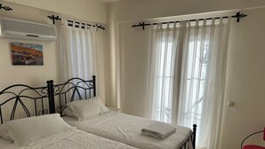A bedroom with two beds, white bedding, a mounted air conditioner, and a painting of a boat on the wall. - 2 bedroom self contained furnished flat with pool, 5 mins walk to town & beach. (Kalkan)