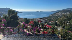 A balcony with vibrant pink flowers, overlooking a coastal town with buildings and a clear blue sky. - 2 bedroom self contained furnished flat with pool, 5 mins walk to town & beach. (Kalkan)