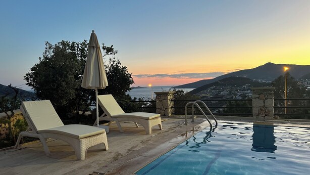 A poolside area with lounge chairs and an umbrella, overlooking a scenic view of a sunset over the sea and hills. - 2 bedroom self contained furnished flat with pool, 5 mins walk to town & beach. (Kalkan)