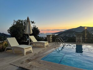 A poolside area with lounge chairs and an umbrella, overlooking a scenic view of a sunset over the sea and hills.