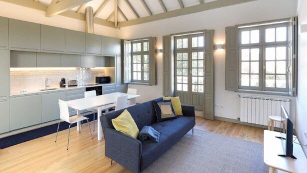 A spacious kitchen with modern appliances, a dining area with a white table and chairs, a grey sofa with cushions, and large windows providing natural light. - Yellow House - The Skylight (Porto)
