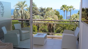 Un balcon doté de meubles en osier, d’une porte vitrée et offrant une vue sur les palmiers et la mer.