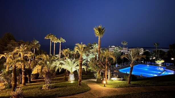 Une vue nocturne d’un complexe hôtelier avec une piscine, des palmiers et un chemin bien éclairé.