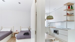 A modern bedroom with two beds, a small bedside table, and a desk with a chair. There is a kitchenette with a microwave and a shelf with items. - Nice and cozy apartment in Pamplona center! (Pamplona/Iruña)