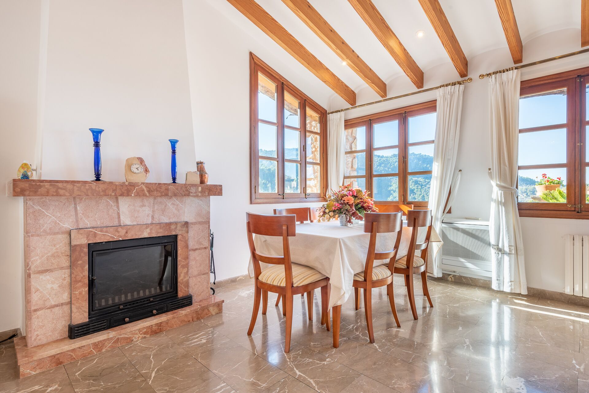 A dining area with a fireplace, wooden ceiling beams, and large windows with curtains.