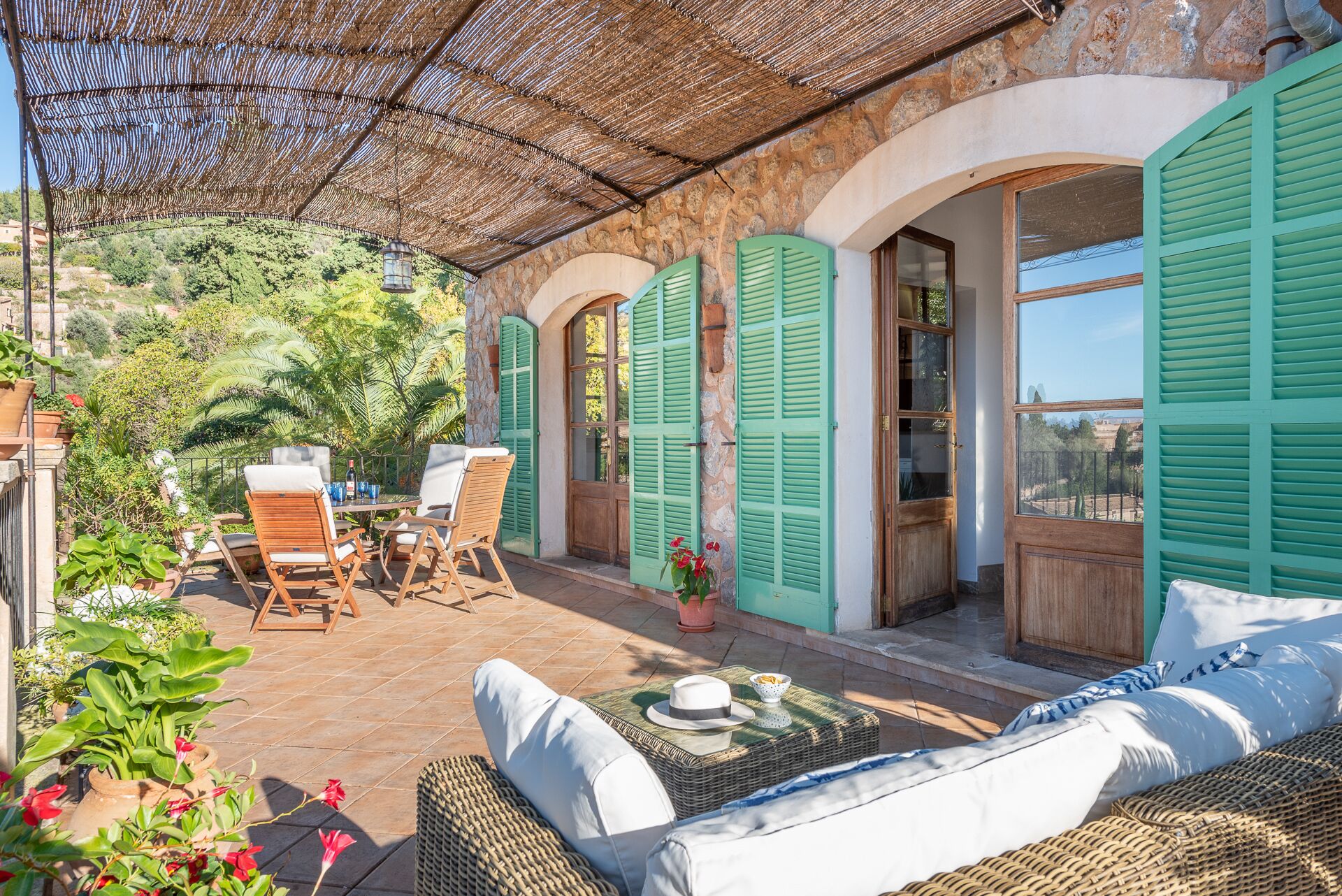 A covered outdoor seating area with wicker furniture, a table with a hat and bowl, and a view of a stone building with green shutters.