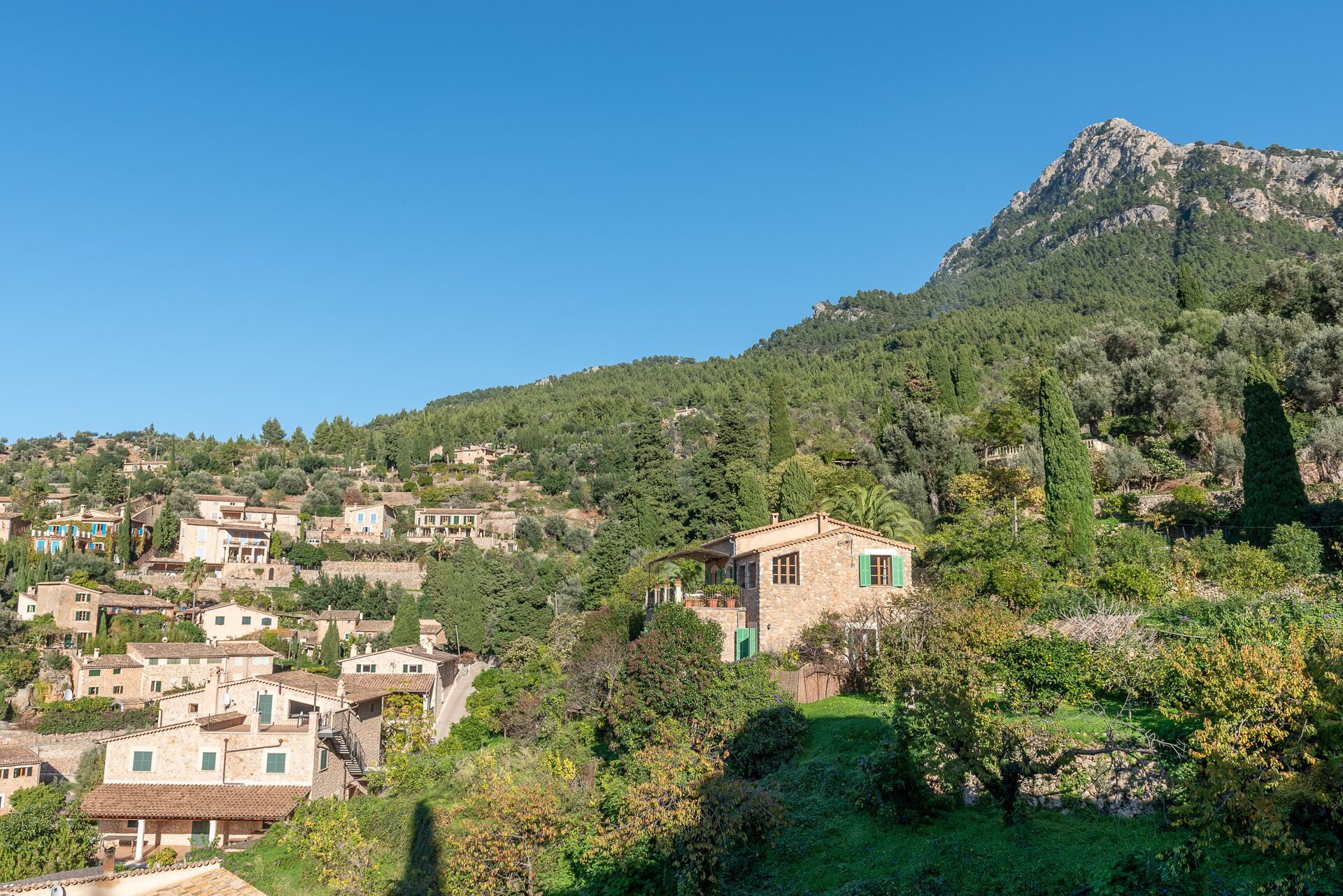 A hillside village with stone buildings, some with green shutters, and a prominent mountain in the background.