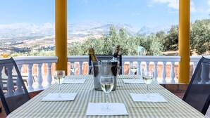 A table set for two with a checkered tablecloth, two wine glasses, and a champagne bottle on a balcony with a mountain view.