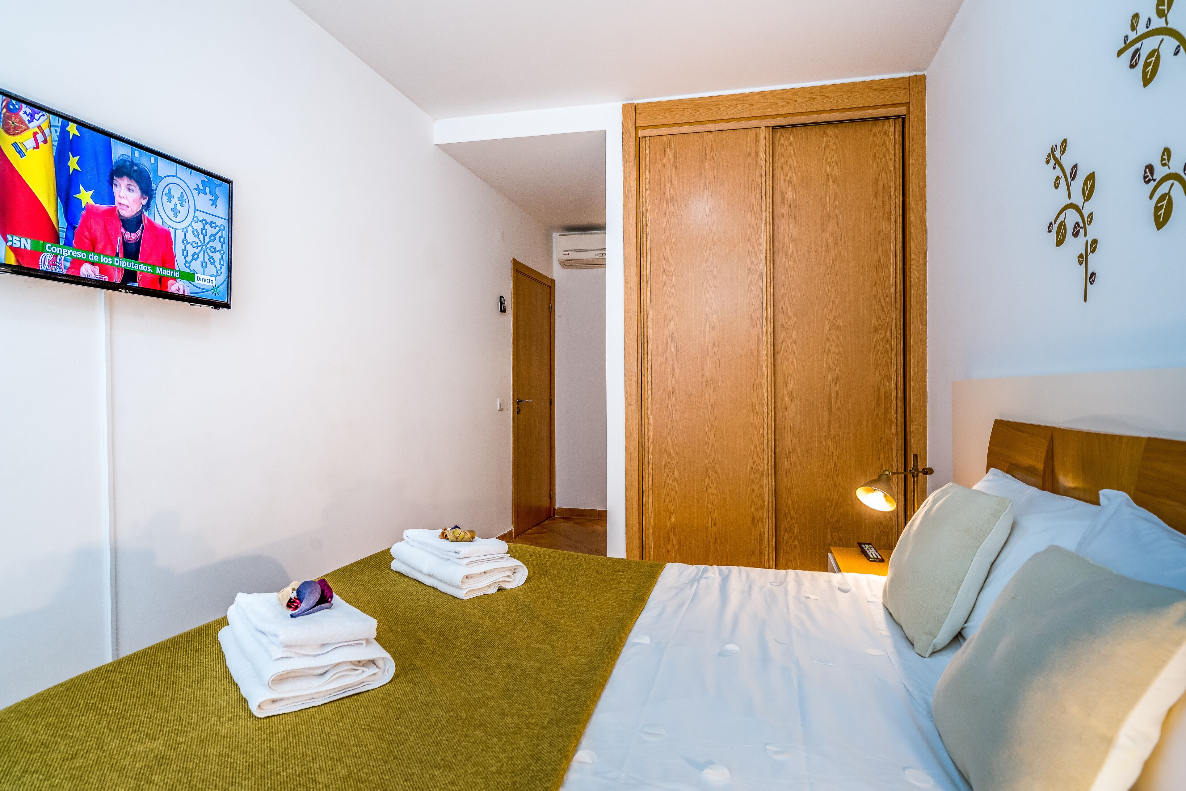 Ein Hotelzimmer mit Bett, Fernseher, der einen Nachrichtensender zeigt, einem Holzschrank und einer Wand mit Dekorationen.
