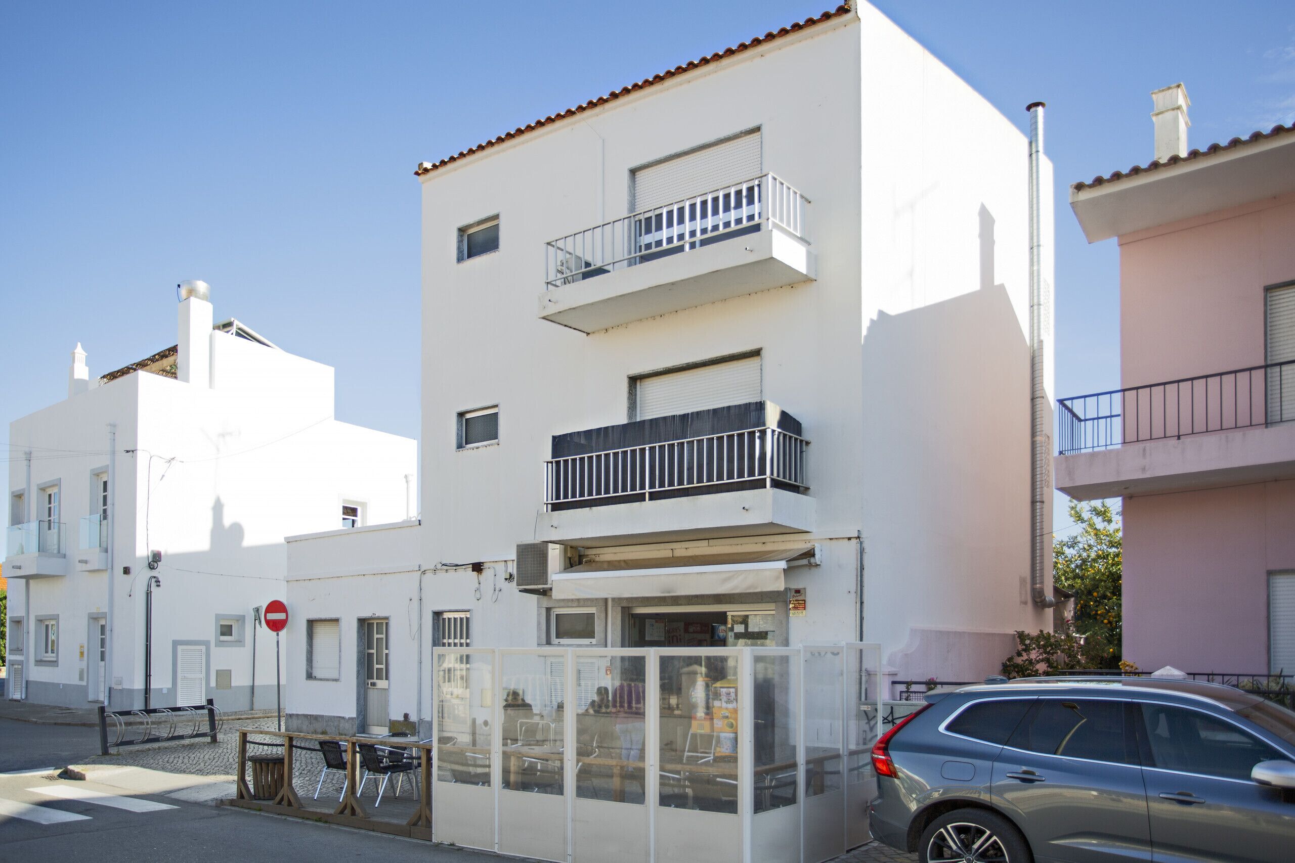 A white building with balconies, a closed shop, and a parked car.