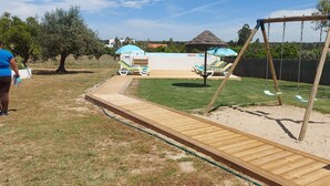 Una terraza de madera que da a un área de juegos de arena con un columpio y sillones de descanso.