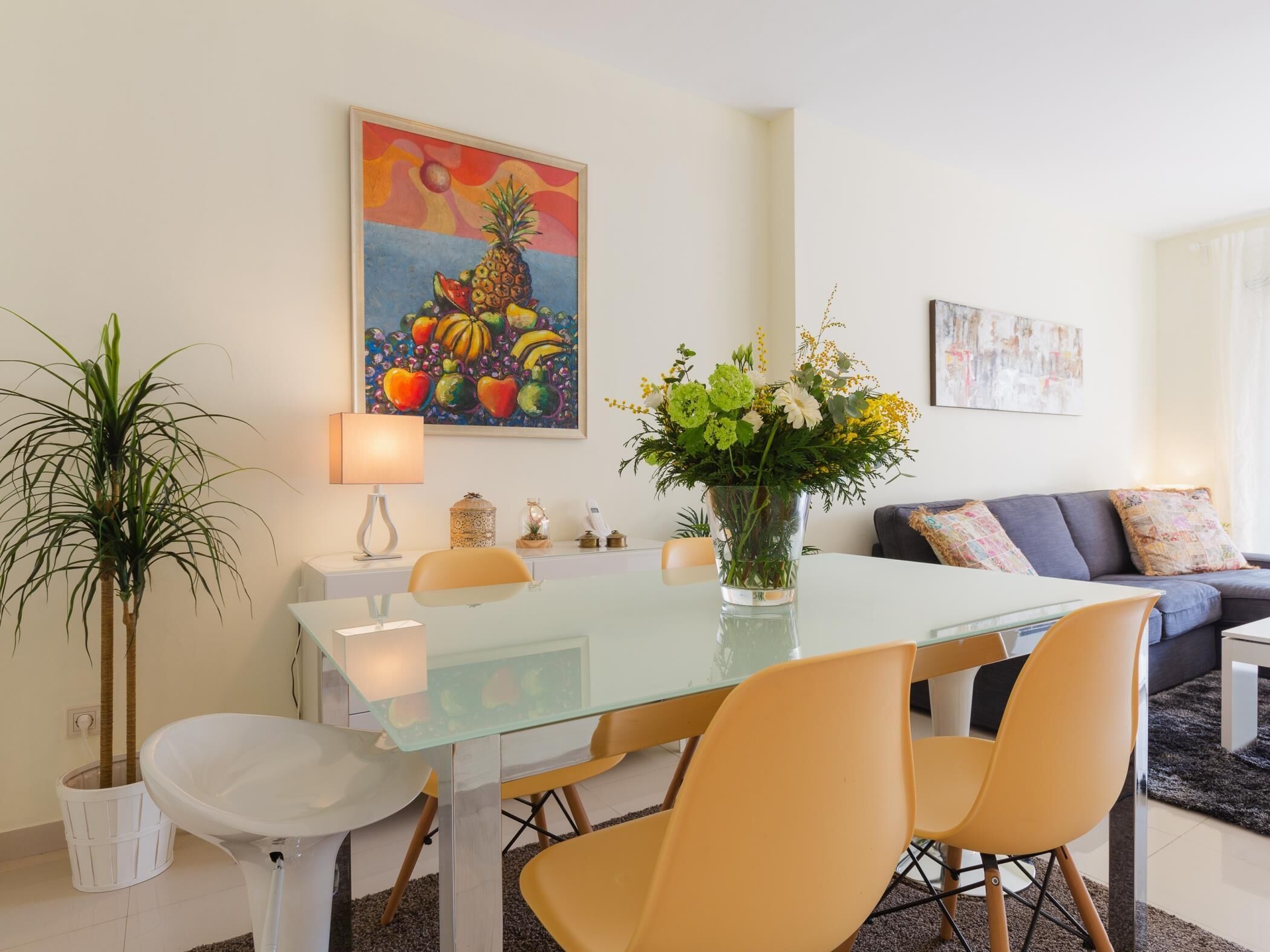 A dining area with a glass table, yellow chairs, a vase of flowers, and a painting of fruits on the wall.