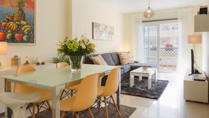 A modern living room with a glass dining table, yellow chairs, a gray sofa, and a coffee table. There is a painting of fruits on the wall and a vase with flowers on the table. - Beautiful apartment near the beach (Rincón de la Victoria)