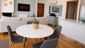 Un saló modern amb un sofà blanc, una taula de dinar rodona amb quatre cadires, un televisor fixat a la paret i una cuina amb un armari blanc i un taulell.