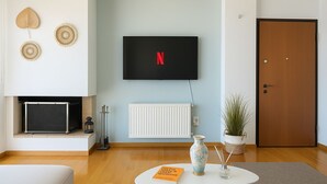 Un saló amb televisió de pantalla plana penjada a la paret, una llar de foc a sota, una taula de cafè blanca amb un llibre i un ram, i una porta a la dreta.