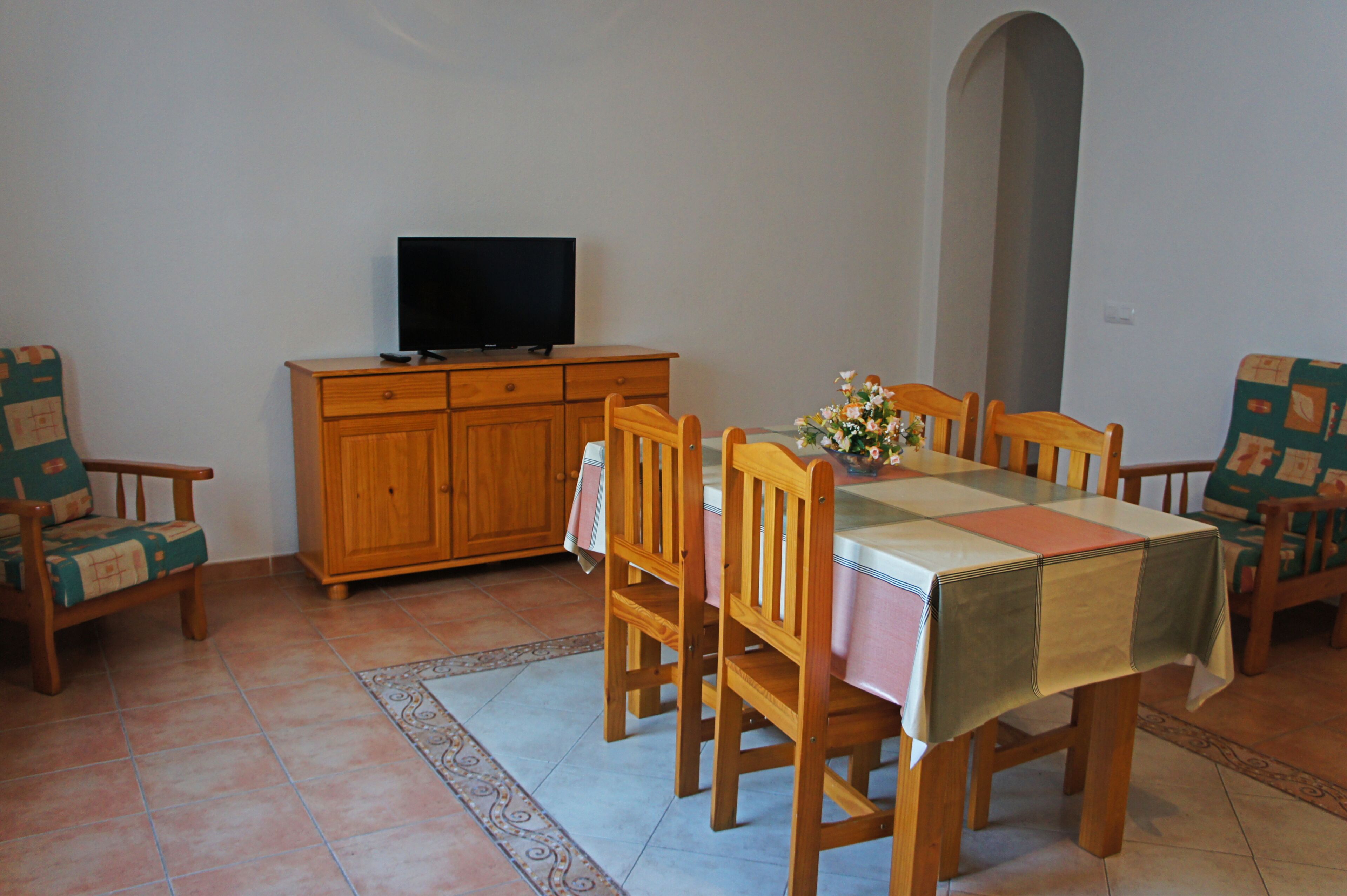 A dining area with a table set for four, wooden chairs, a television on a cabinet, and a patterned rug on the floor.