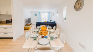 Un comedor moderno y bien iluminado, con una mesa de madera dispuesta para cuatro personas, rodeada de sillas blancas. Un reloj grande está colgado en la pared, y se ve un sofá azul al fondo.