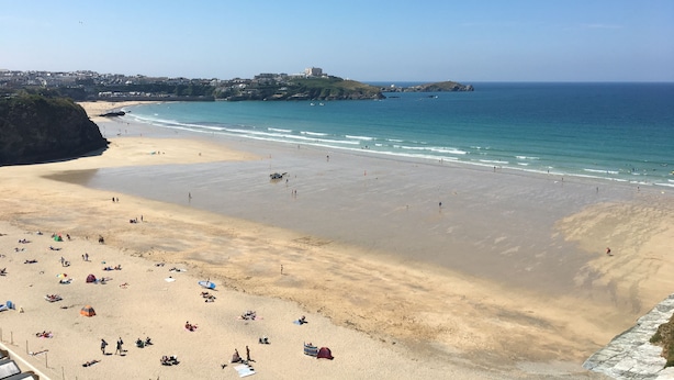 Una amplia playa de arena con gente y sombrillas, cielo azul despejado y una vista distante de un pueblo y la costa.