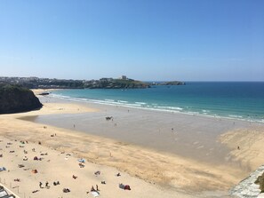 Una amplia playa de arena con gente y sombrillas, cielo azul despejado y una vista distante de un pueblo y la costa.