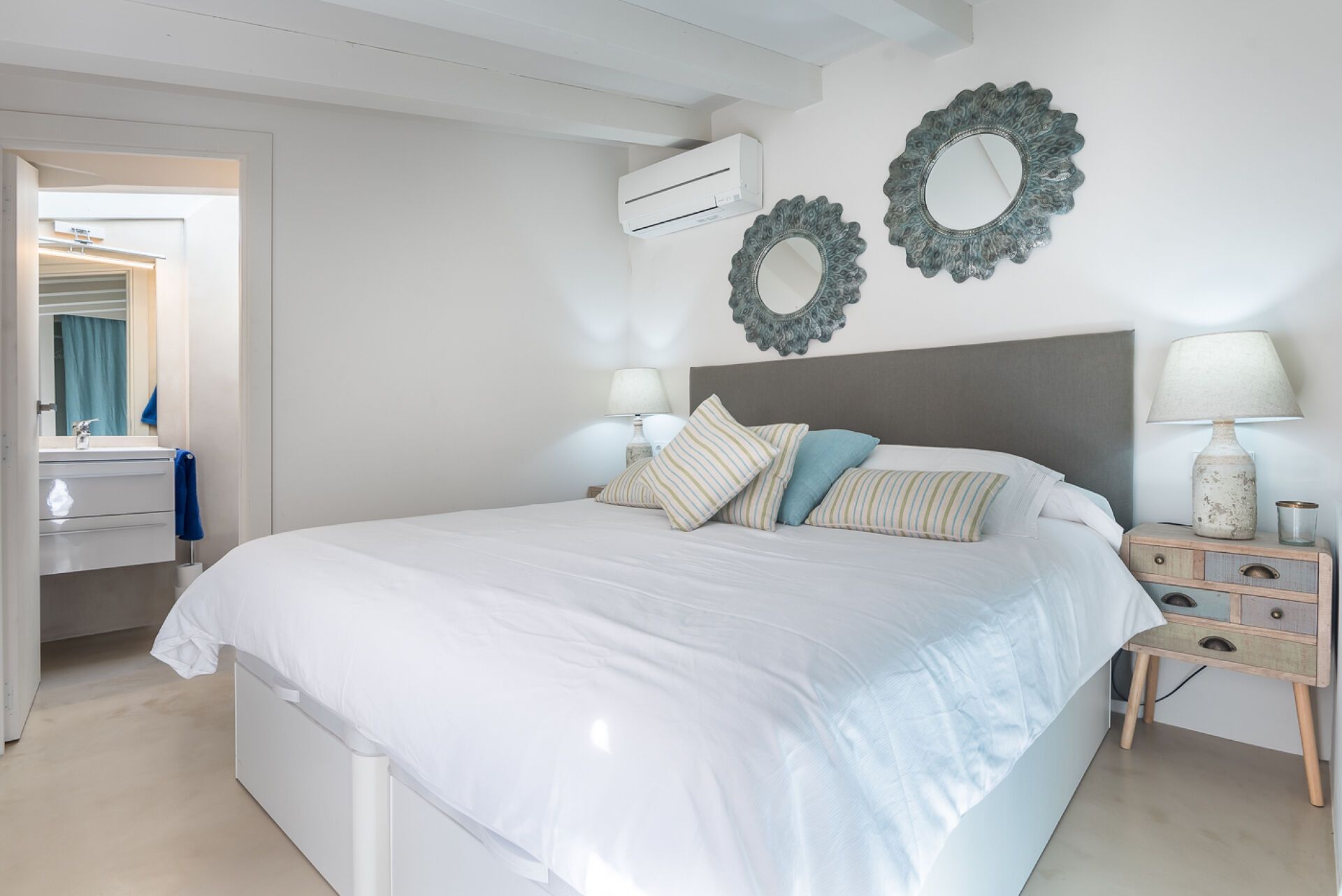 A bedroom with a large bed, two wall mirrors, a bedside table with a lamp, and an air conditioning unit.
