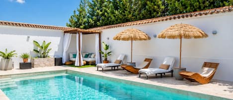 Un área de piscina con sillones de descanso, una pérgola y plantas en macetas.