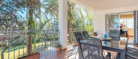 Un balcón con una mesa y sillas de vidrio, con vistas a un jardín con palmeras y el cielo.