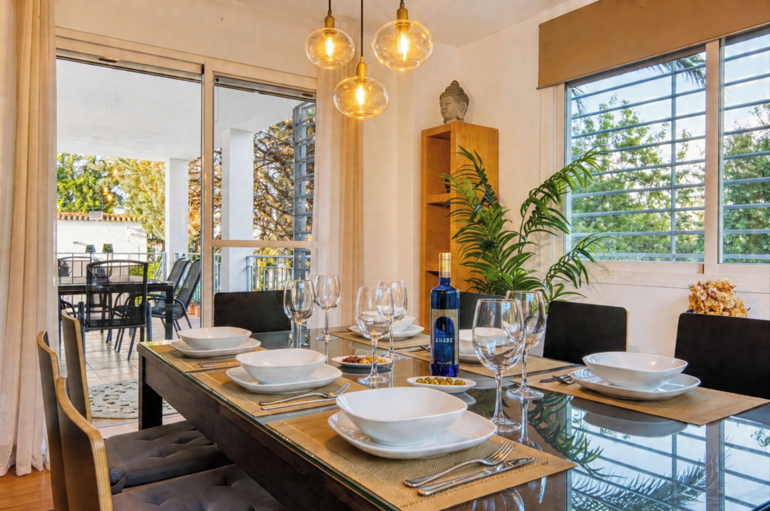 Un coin repas avec une table en verre dressée pour un repas, entourée de chaises, et en arrière-plan, une statue de Bouddha et une plante.