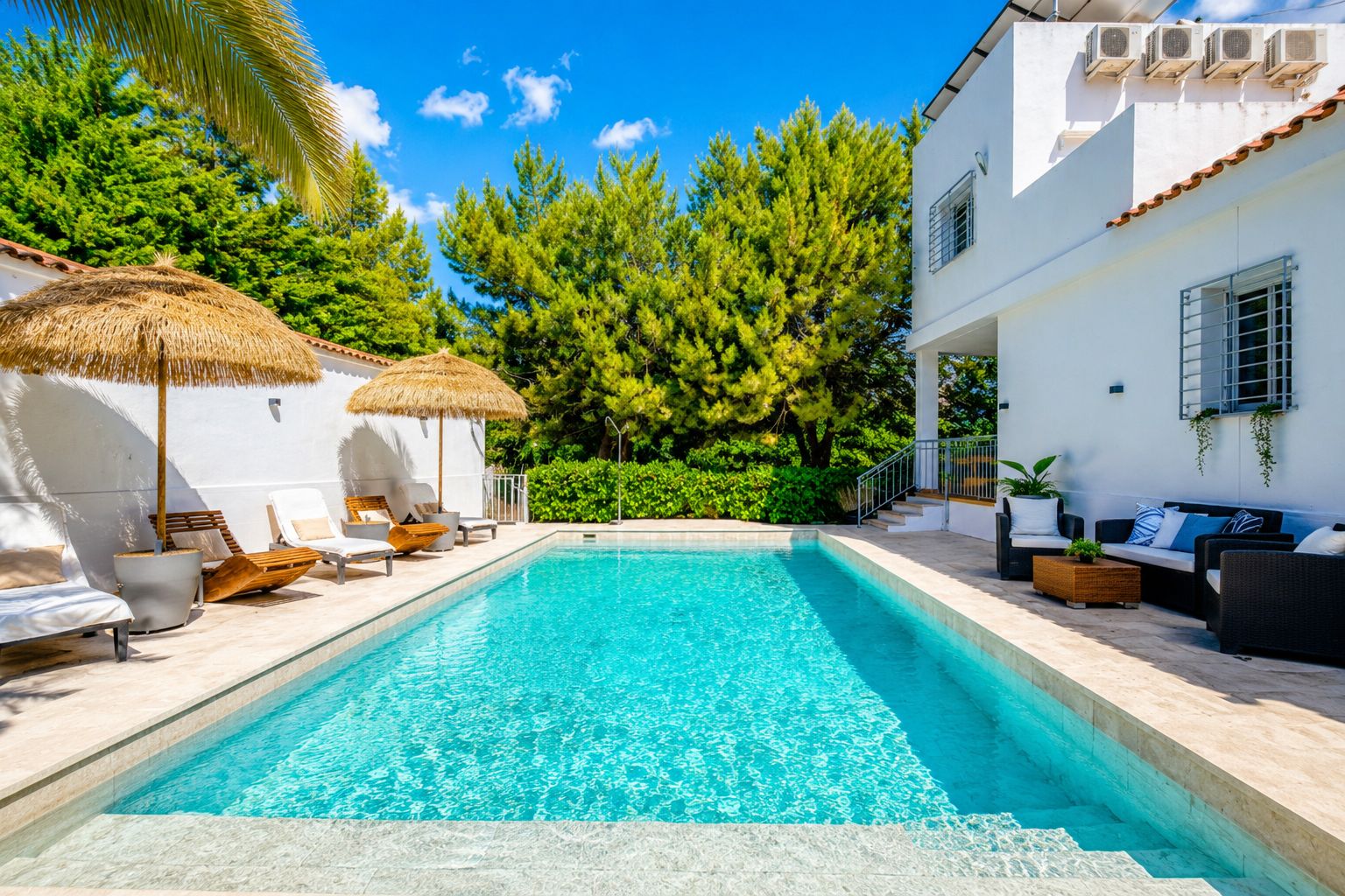 Una piscina amb gandules i ombrelles, envoltada de vegetació i un edifici blanc.