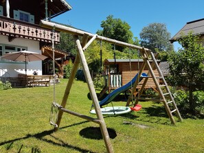A wooden swing set and slide in a backyard with a dining area and a house in the background.